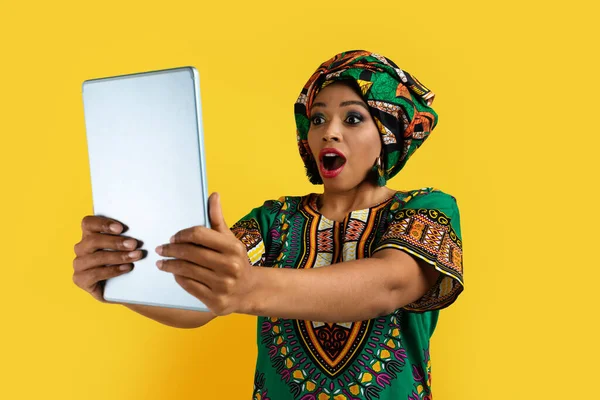 令人难以置信的网上交易 穿着传统服装 看着现代数码显示屏 高喊着黄色工作室背景的年轻非洲姑娘惊愕不已 — 图库照片