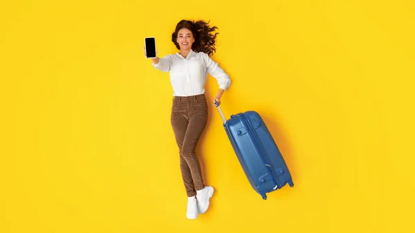 旅行の申し込み 携帯電話のブランク画面を表示するハッピーレディ観光客は黄色のスタジオの背景にポーズスーツケース 広告アプリケーションや予約旅行チケットを保持します パノラマ — ストック写真
