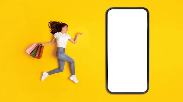 stock image Cheerful Lady Buyer With Shopping Bags Jumping Near Large Smartphone With Blank Screen, Advertising Ecommerce Offer Or Application On Yellow Studio Background. Great Sale Concept. Panorama, Mockup