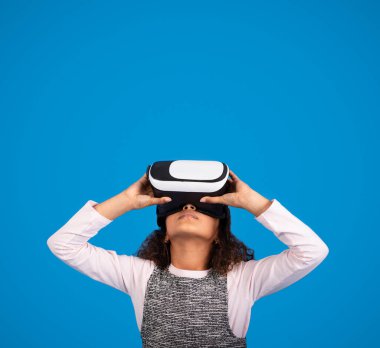 VR gözlüklü siyah bir kız çevrimiçi oyun oynuyor, eğleniyor, boş alana bakıyor, mavi arka planda, stüdyoda izole halde. Reklam ve teklif, aygıt ile eğitim, modern teknoloji ve çocukluk