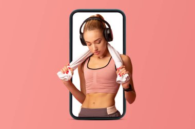 Spor giyimli yorgun Avrupalı bir bayan ve pembe stüdyo arka planında izole edilmiş akıllı telefon ekranında dinlenen kulaklıklı bir kulaklık. Spor, kilo verme, vücut bakımı için müzik uygulaması