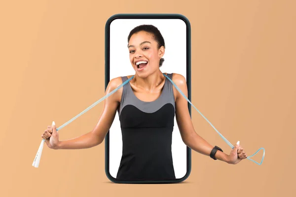 Happy young black lady in sportswear with open mouth and jump rope on phone screen isolated on beige studio background, collage. Fitness, workout, weight loss and health care