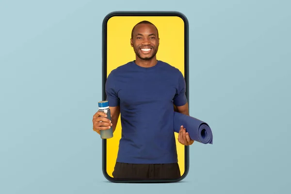 青のスタジオの背景に隔離されたスマートフォンの画面上のマットと水のボトルでスポーツウェアで陽気な若いアフリカ系アメリカ人の男運動選手のコーチ カクテル ボディケア ワークアウトアプリでフィット — ストック写真
