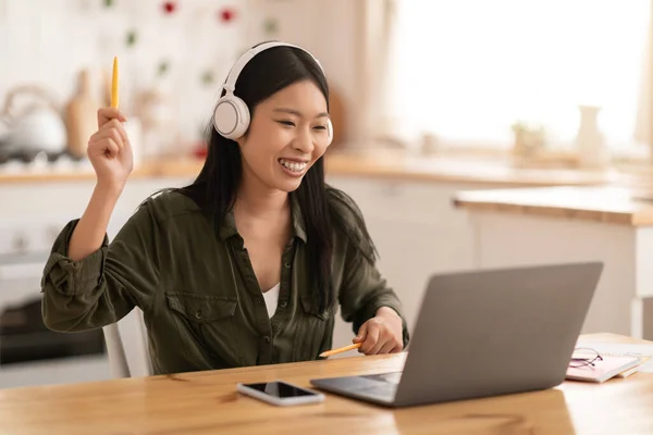 Joyful young asian woman having break while working from home, sitting at kitchen table, holding pencils, using wireless stereo headphones and modern laptop, listening to music, copy space