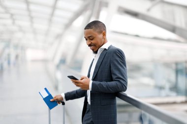 Online Check-In. Havaalanında uçuşu beklerken akıllı telefon kullanan gülümseyen siyahi işadamı, Terminal Salonu 'nda beklerken İnternet' i tarayan takım elbiseli yakışıklı Afrikalı Amerikalı erkek.