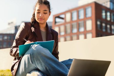 Çevrimiçi eğitim kavramı. Bayan karışık ırk öğrencisi dışarıda dizüstü bilgisayarla çalışıyor, kadın şehir parkında oturuyor ve not defterine not alıyor, uzaktan ders alıyor.