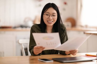 Gözlüklü neşeli genç Asyalı kadın mutfak masasında dizüstü bilgisayar ve telefonla oturuyor, elinde kağıtlar, mektupları kontrol ediyor ya da evden gelen belgelerle çalışıyor.