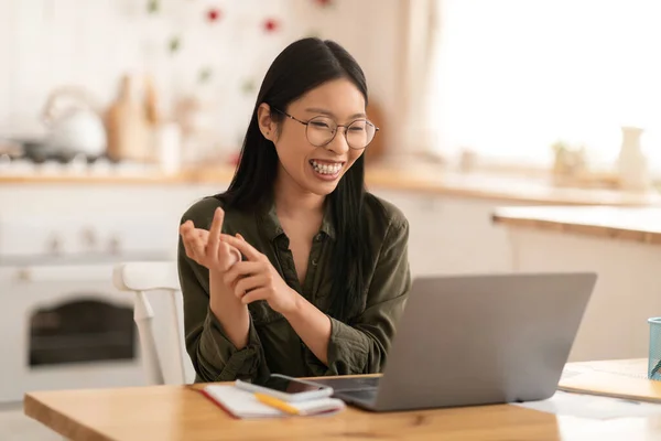 Happy pretty young chinese woman in casual sitting at kitchen table, have online meeting with business partners, using laptop, looking at computer screen, smiling and gesturing, copy space