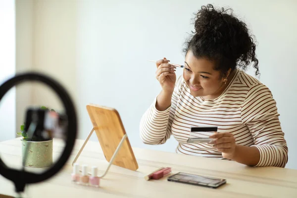 Beauty blog, blogging concept. Cheerful pretty curly young woman plus size in casual outfit sitting at desk in front of mirror at home, recording video while applying makeup, using blogger set