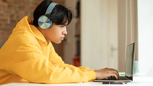 オンライン研究 ノートパソコンでサーフィンをしたり 宿題をしたり 自宅でヘッドフォンをしている日本人学生 現代の距離教育と家庭教育の概念 サイドビューショット パノラマ — ストック写真