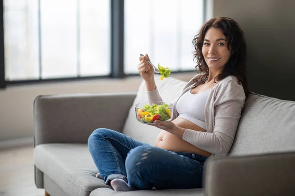 妊娠中の栄養ニーズ 妊娠中の女性は自宅で新鮮な野菜サラダを食べます 笑顔若い期待の母親は ソファに座って 健康的な食事を楽しみます ビタミン食品を持っています スペースをコピー — ストック写真