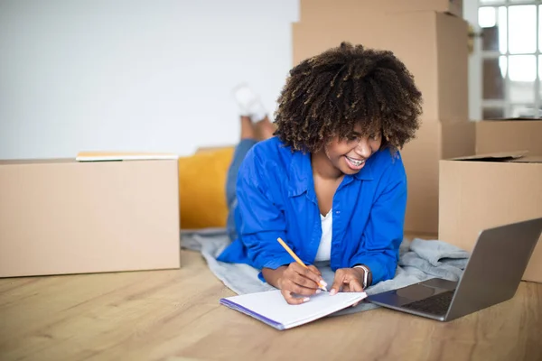 Smiling Black Woman Using Laptop And Making Checklist While Moving Home, Young African American Female Browsing Internet On Computer And Writing In Notepad While Resting Among Cardboard Boxes
