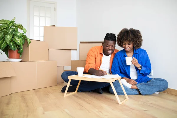 Young Black Family Couple Planning Shopping For New Home, Making Checklist While Sitting Among Cardboard Boxes In Their Flat, African American Spouses Drinking Coffee And Writing In Notepad
