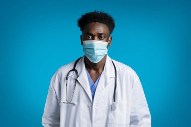 Boynunda steteskop olan, beyaz ceketli yakışıklı zenci bir doktorun portresi. Doktor yüz maskesi takıyor. Mavi stüdyo arka planında izole edilmiş.