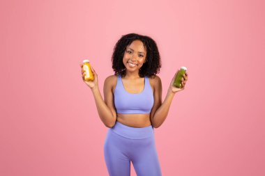 Binlerce yıllık Afro-Amerikan kadın spor kıyafetleri içinde meyve suyu ya da meyve suyu kavanozları tutuyor, sağlıklı içeceklerin tadını çıkarıyor, pembe arka planda, stüdyoda izole edilmiş. Diyet, spor vitaminleri, reklam ve teklif, vücut bakımı