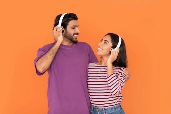 一緒に音楽をお楽しみください 幸せな笑みを浮かべて若いインドのカップルは ワイヤレスヘッドフォンを使用して 歌を聞いて 抱き合って見て オレンジの背景に隔離された スタジオショット — ストック写真
