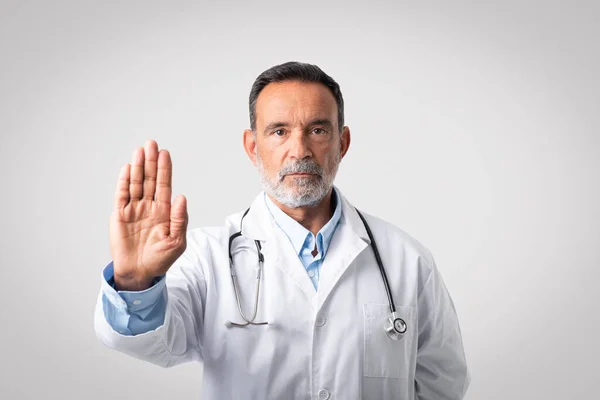 重大な自信を持ったヨーロッパの先輩医師が聴診器で白衣を着てグレーの背景に手でサインを止めます 専門的な治療 医療との戦い — ストック写真