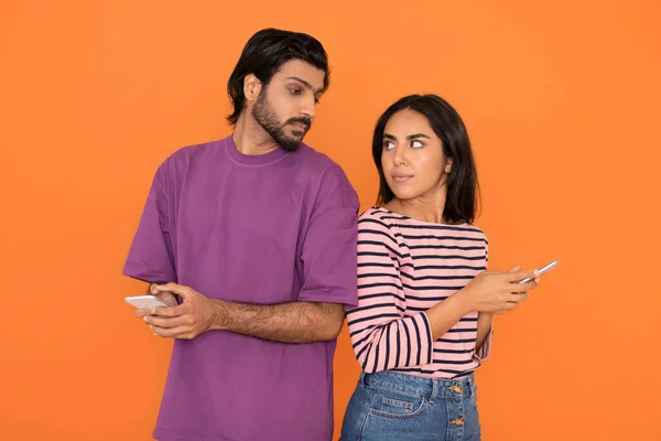 関係の概念で浮気や不貞 若いインド人男性が女性に背を向け電話でメッセージを読みスクリーンを覗こうとしたオレンジのスタジオの背景 — ストック写真