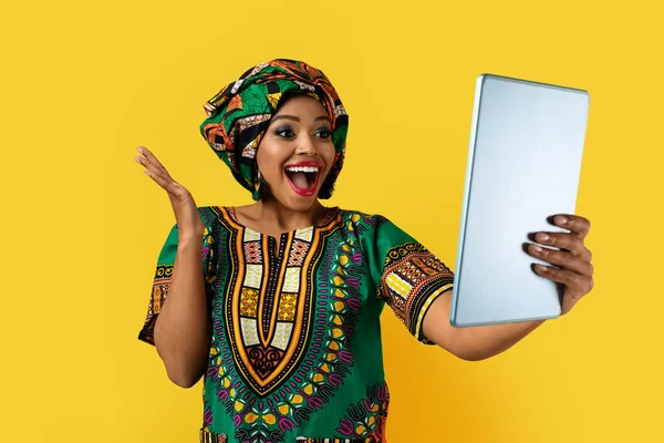 快乐而又漂亮的年轻黑人女性 身穿传统的非洲服装 手握现代数码平板电脑 头脑清醒 在黄色的工作室背景下与外界隔绝 — 图库照片