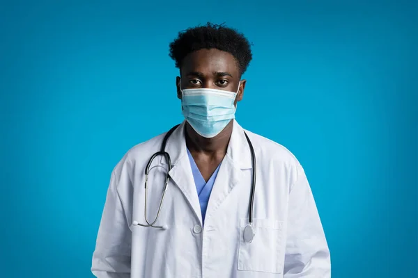 若いハンサムな黒人医師の肖像画白い医療コートを着て 首に聴診器 顔のマスクを身に着けている医者 青いスタジオの背景に隔離された コピースペース — ストック写真