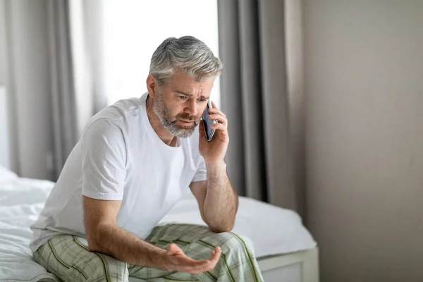 怒って感情的なハンサムな中年の男性のベッドの上に自宅で座って 携帯電話の会話をして 恋人やビジネスパートナーとの戦い パジャマを着て スペースをコピーします コミュニケーションの概念 — ストック写真