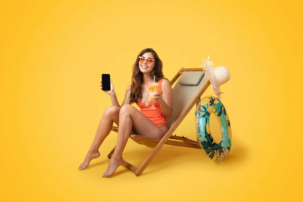 水着を着た幸せな千年のアジアの女性 デッキチェアに座ってインフレータブルリングを持つサングラス 熱帯カクテル 休息をお楽しみください オレンジのスタジオの背景に隔離された空白の画面でスマートフォンを表示 — ストック写真