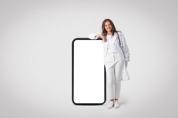 Telehealth platform partnerships. Senior doctor woman in uniform standing near huge blank smartphone, demonstrating space for design, app or website, mockup