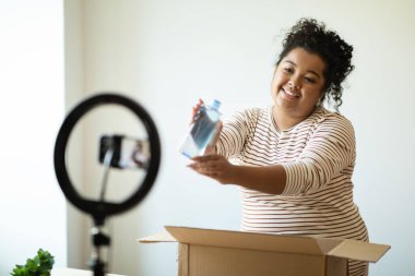Güzellik blogcusu, tatlı, genç, İspanyol kıvırcık kadın artı evde paketleri açan, blog 'u için video kaydeden, cep telefonu ve tripod kullanan, mikiler suyu, toner gösteren.