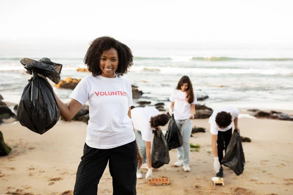 高兴的是 年轻的国际志愿者穿着T恤衫 非洲裔美国妇女背着垃圾袋 在海滩上清理垃圾 生态和环境保护 地球日户外 — 图库照片