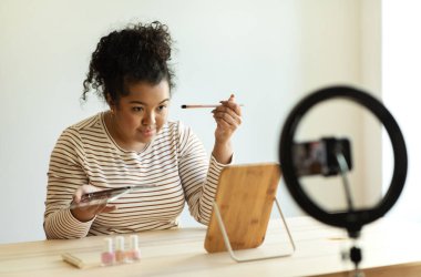 Güzel gülümseyen kıvırcık İspanyol genç bayan artı boyut makyaj sanatçısı akıllı telefonuyla güzellik blogu için video kaydediyor. Masasında oturan kadın, yepyeni güzellik ürünlerini test ediyor, göz falı bakıyor.