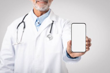 Steteskoplu beyaz önlüklü gülümseyen Avrupalı kıdemli doktor gri arka planda boş ekranlı akıllı telefon gösteriyor. Profesyonel danışmanlık ve tıbbi bakım, blog, web sitesi, reklam ve teklif
