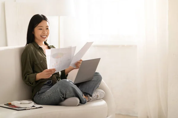 幸せな若いアジアの女性のビジネスアナリスト 会計士は彼女の膝の上にラップトップでソファに座って 紙を保持し 対応をチェックしたり 自宅から文書で作業したり スペースをコピーします — ストック写真