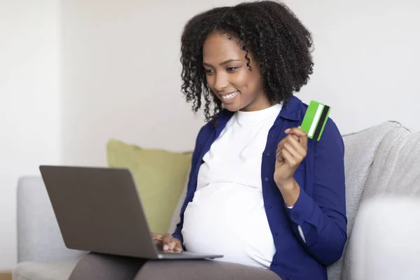 ラップトップ付きの大きな腹を持つ陽気な千年のアフリカ系アメリカ人の妊婦は リビングルームのインテリアでソファの上にクレジットカードを示しています 赤ちゃん オンラインショッピング 巨大な販売とキャッシュバックを期待 — ストック写真