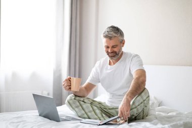 Evde ya da otel odasında pijama giyen, sabahları kahve içen, dizüstü bilgisayarını kullanan, ajandasını kontrol eden mutlu, orta yaşlı iş adamı.