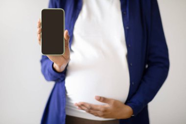 Milenyum Afrika kökenli hamile kadın oturma odasının içinde boş ekranlı akıllı telefon gösteriyor. Bebek bekleniyor, iletişim uygulaması, web sitesi tavsiyesi, blog