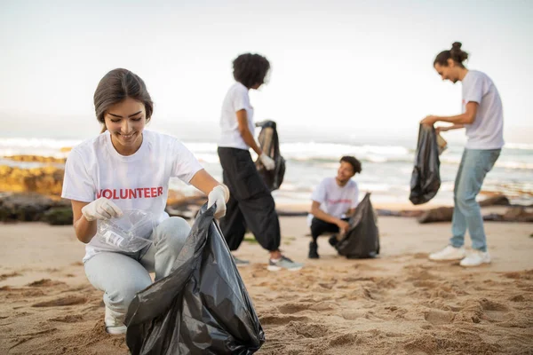 积极的国际青年志愿者戴着手套 欧洲裔女士背着垃圾袋清理海滩上的垃圾 保护地球的生态和环境保护 海洋污染 — 图库照片