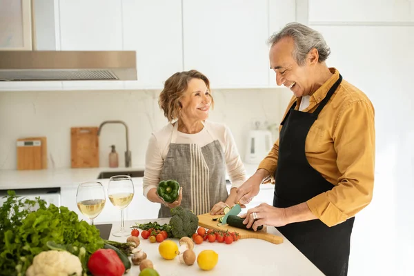 Happy elderly spouses cooking together and talking, woman and man preparing vegetable salad, making dinner together, kitchen interior, copy space