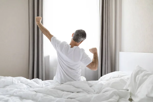 目に見えない灰色の髪の男のパジャマのベッドの上に座って 午前中に目を覚ました後 体を伸ばし ウィンドウを見て コピースペースのバックビュー 快適な健康的な睡眠コンセプト — ストック写真
