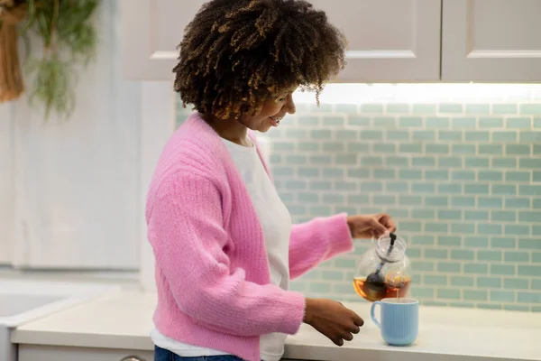 stock image Side view of happy young stylish black lady wearing casual outfit brewing tea at kitchen, enjoying relaxing aromatic herbal tea after working day, copy space. Domestic lifestyle