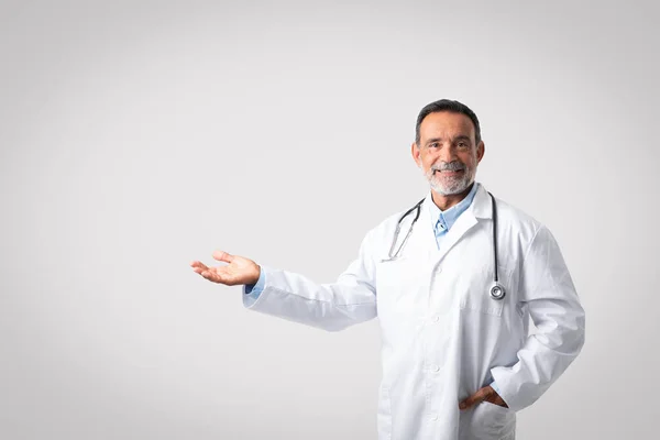 聴診器付き白衣を着た陽気で自信のあるヨーロッパの上級医師は 灰色の背景に無料のスペースを持つ手を示しています クリニックでの診断 専門家のアドバイス 広告とオファー — ストック写真