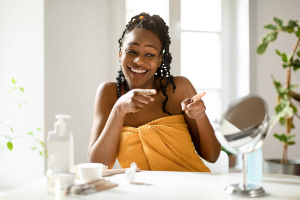 Power of daily skincare. Happy african american lady pointing fingers at her reflection in small mirror, sitting at dressing table in bedroom while making daily beauty routine