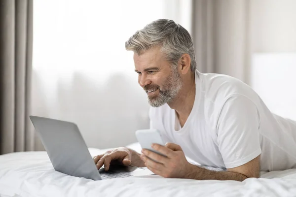 快乐英俊的白发中年男子数字游牧民族穿着睡衣躺在床上 手持手机和笔记本电脑 侧视复制空间 现代技术概念 — 图库照片