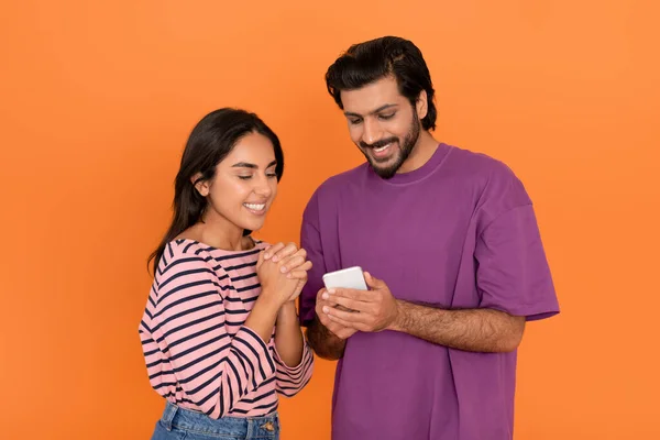 宝くじ キャッシュバック プレゼント 正の幸せなかわいい若いインドのカップルを使用して携帯電話一緒に 笑顔興奮した東部の恋人ギャンブルインターネット上で オレンジのスタジオの背景 — ストック写真