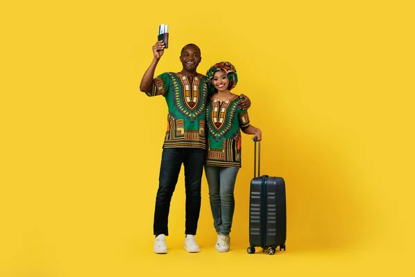 黄色のスタジオの背景を採用し 荷物を運んで 航空券やパスポートを示す伝統的なカラフルなドレッシングで幸せな興奮した美しいアフリカのカップル — ストック写真