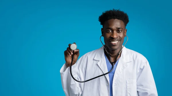 青いスタジオの背景に白い医療コートの医師を身に着けているハンサムな陽気なフレンドリーな若いアフリカ系アメリカ人男性は 聴診器と笑顔を使用して コピースペース パノラマ 仕事と職業 — ストック写真