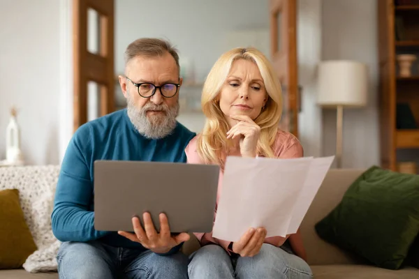 Balancing the Budget. Serious Mature Spouses Thoughtfully Reviewing Paper Bills And Using Laptop, Managing Finances Online Sitting On Sofa At Home. Household Paperwork And Retirement Concept