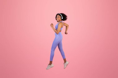 Bin yıllık Afro-Amerikan bayan spor giyim, kablosuz kulaklıklar müzik, dans, zıplama, pembe arka planda ve stüdyoda donma. Spor, reklam ve teklif için ses uygulaması