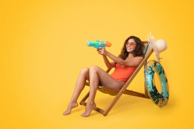 İyi ki bin yıllık Asyalı mayo giymiş, güneş gözlüklü bir kadın güverte sandalyesine oturmuş, boş zamanın tadını çıkarıyor ve turuncu stüdyo arka planında izole edilmiş su tabancası oynuyor. Yaz eğlencesi, dinlenme ve dinlenme, boş zaman