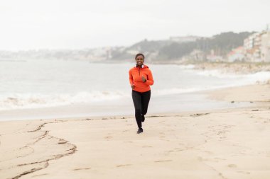 Spor kıyafetleri ve kulaklıklar takan mutlu Afro-Amerikan kadın deniz sahilinde koşuyor, dışarıda antrenmanların keyfini çıkarıyor, tam boy. Vücut bakımı, tek başına eğitim, aktif yaşam tarzı ve kilo kaybı