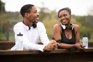Spor giyim kuşamında gülümseyen Afro-Amerikan bir çift. Su şişeli kulaklıklar açık hava parkında koştuktan sonra dinleniyor. Kilo kaybı, vücut bakımı, şehirde çalışma, birlikte ayrılma.